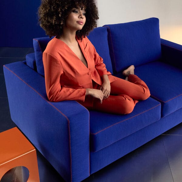 Innovation Living Neah X -vuodesohva, sininen Elegance Ultramarine -kangasverhoilu. Miljöökuvassa nainen istuu sohvalla oranssissa asussa.