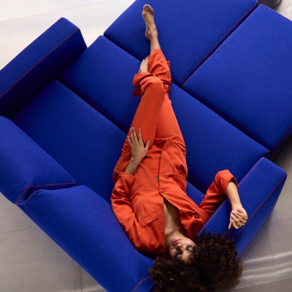 Innovation Living Neah X -vuodesohva, sininen Elegance Ultramarine -kangasverhoilu. Miljöökuvassa nainen lepää sohvalla oranssissa asussa.