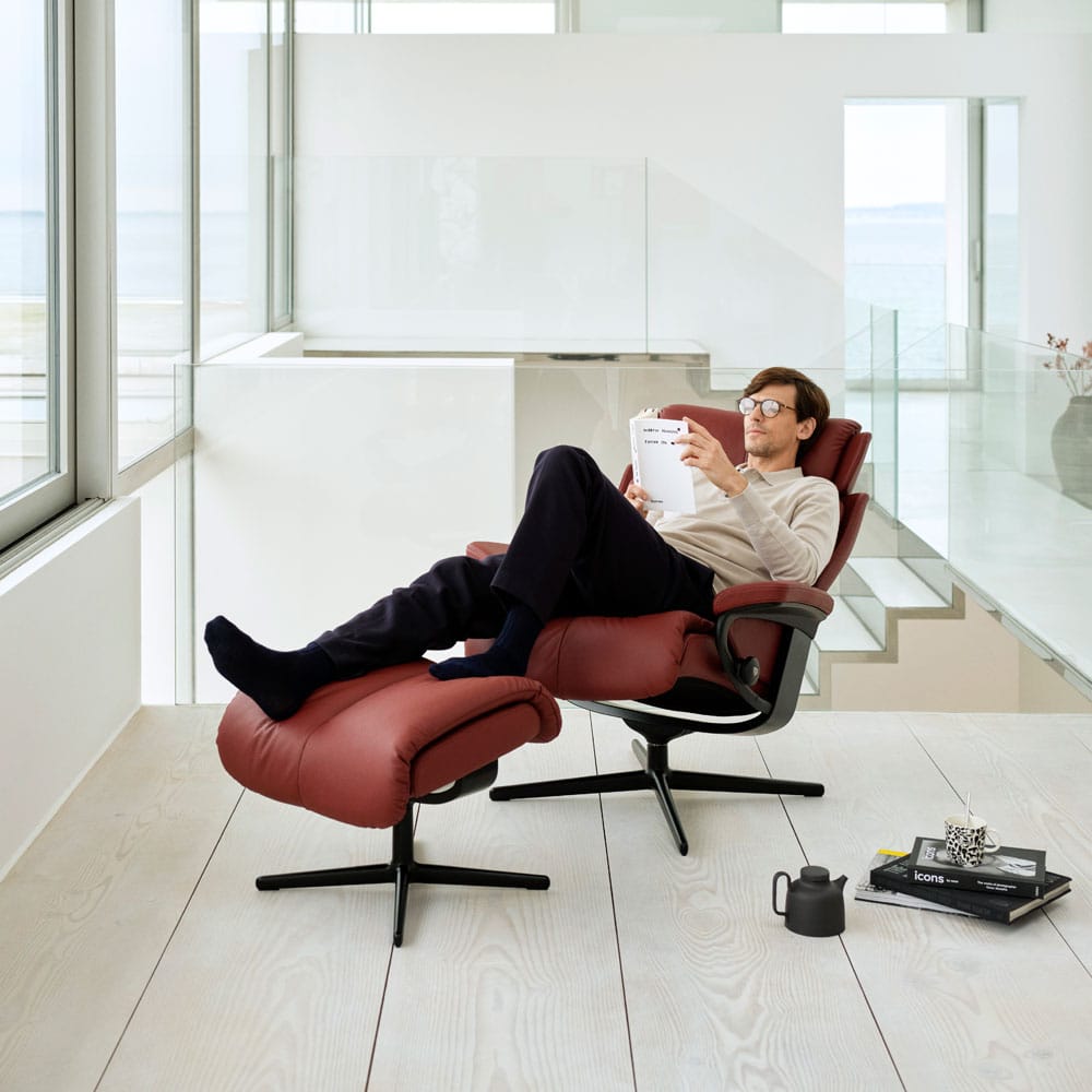 Stressless Magic-lepotuoli. Mijöökuvan tuolissa on punainen nahkaverhoilu ja musta Cross-jalka. Mies lepää tuolilla lukien kirjaa.